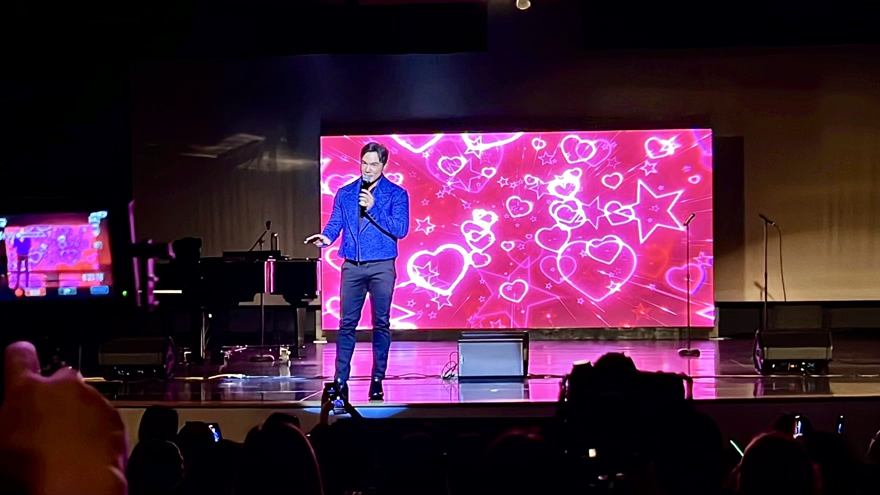 Mark Bautista is on stage singing a love song to the audience. A vibrant large LED display filled with hearts is behind him.