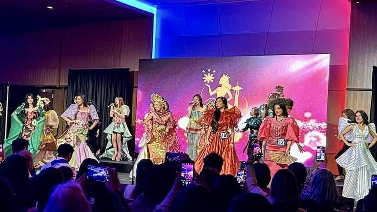 Several contestants in a beauty pageant stand on stage, each wearing their national costume. A large LED display behind them shows the pageant logo over a colorful abstract pattern.